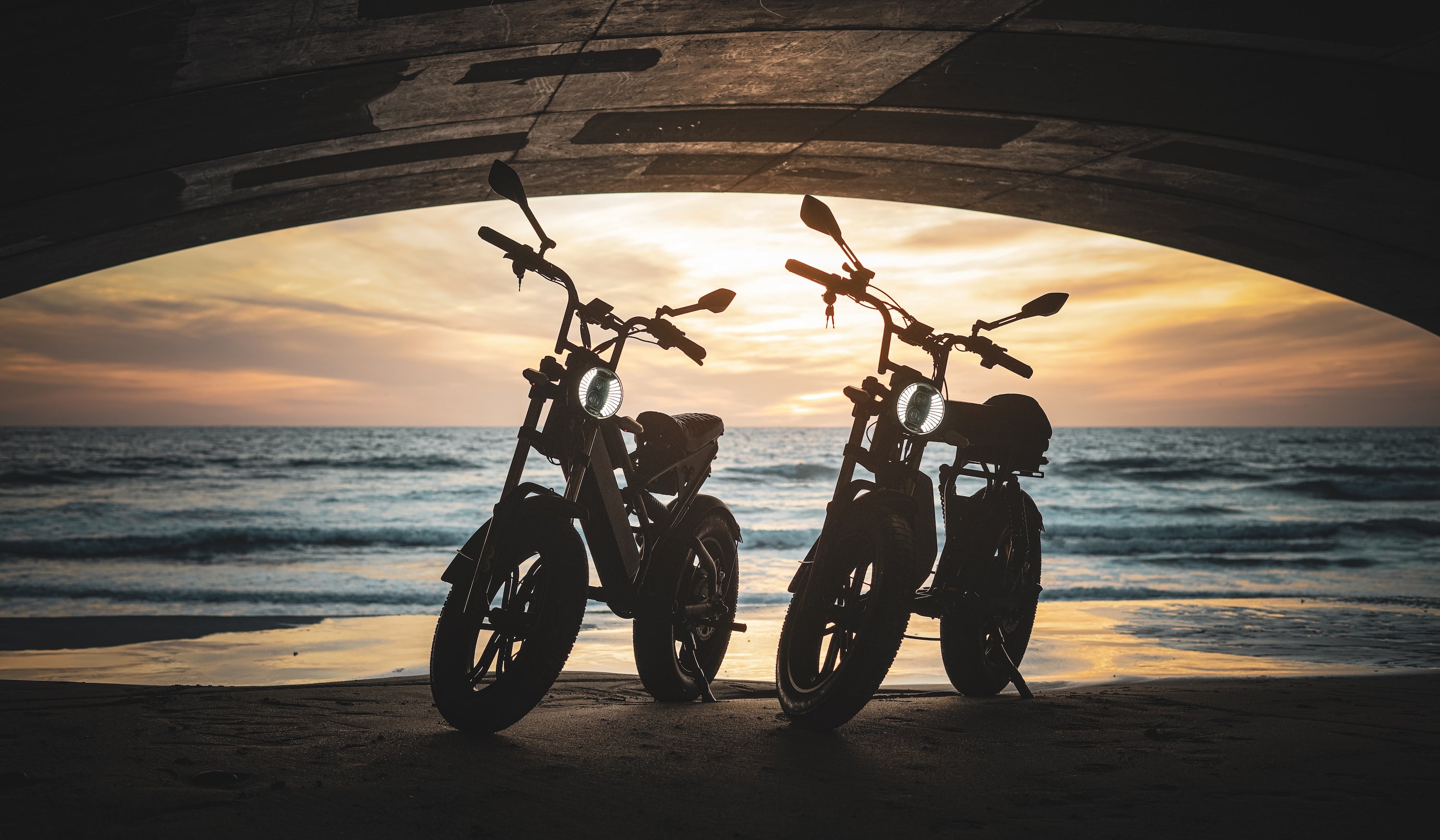 Two motorcycles silhouetted against a sunset over the ocean.
