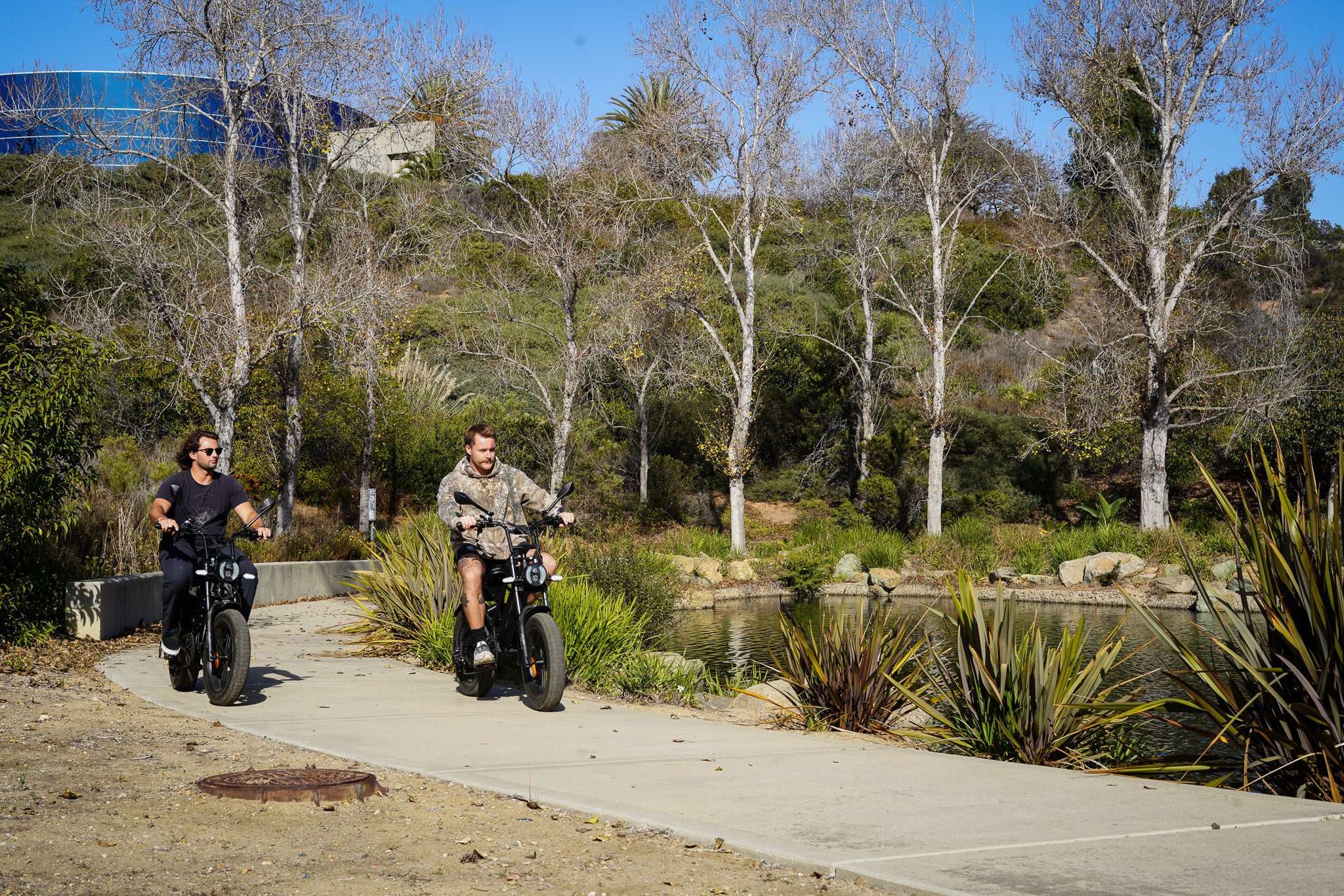 Two people riding electric bikes on a path with trees and a building in the background
